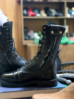 Load image into Gallery viewer, Black leather military boots by leather gear on a table with a background of shoes on shelves.
